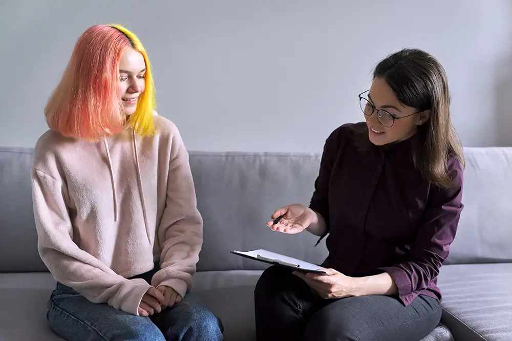 A young adult sitting on a couch talking to a female with a clipboard