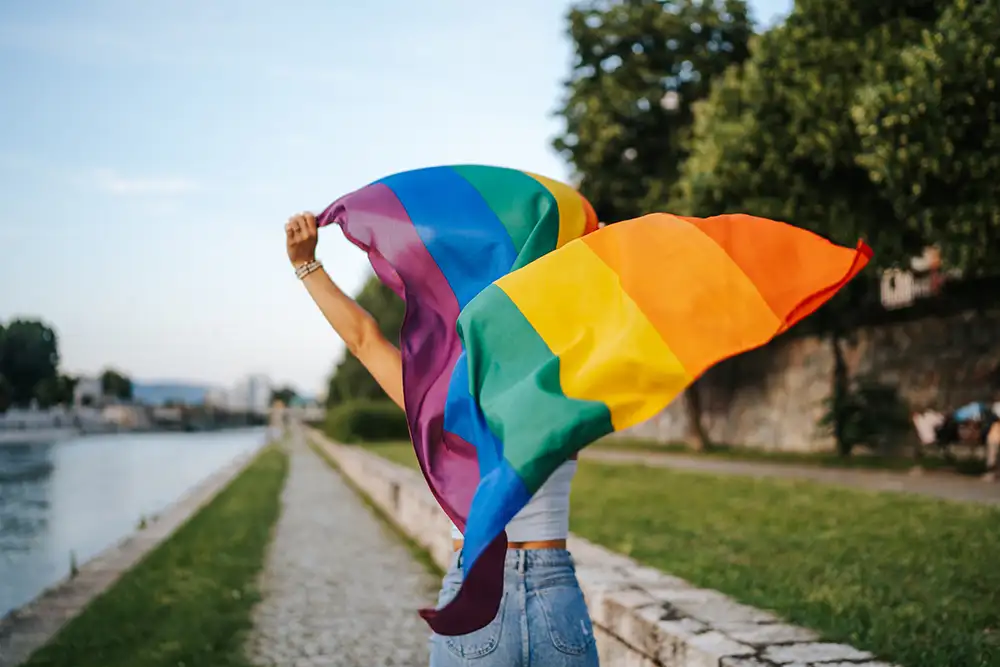 A person holding a rainbow flag above their head while walking along a river