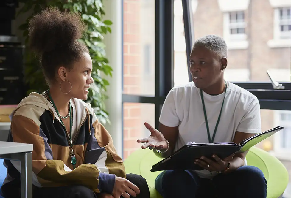 Two professionals talking to each other infront of a window while one holds a clipboard